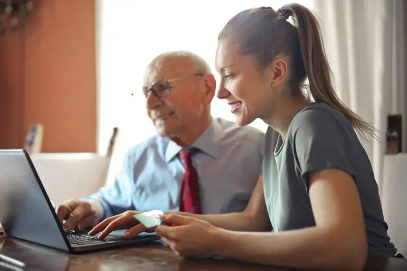 Elderly grandfather and young granddaughter sit side-by-side at a laptop, both smiling while she points at the screen and he rests a gentle hand on her shoulder.